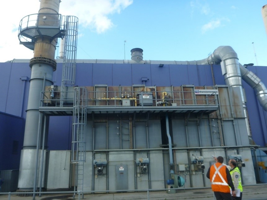 Photo of a large scale industrial thermal oxidiser. Two people wearing high-vis vests are standing in front of it, in the bottom right corner of the photo.